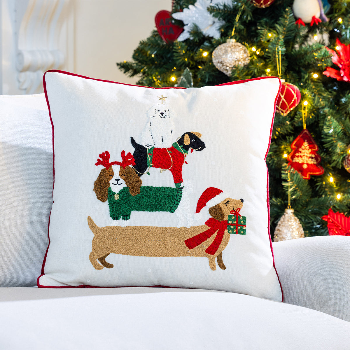 White Christmas cushion featuring four festive dogs stacked together, displayed on a white sofa beside a decorated Christmas tree.