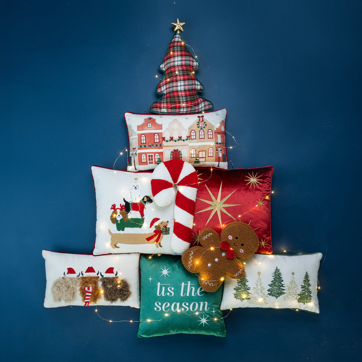 A collection of festive Christmas cushions arranged with fairy lights against a dark blue background.