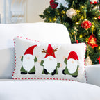 White Christmas cushion featuring three embroidered gnomes in green outfits and red hats, displayed on a white sofa beside a Christmas tree.