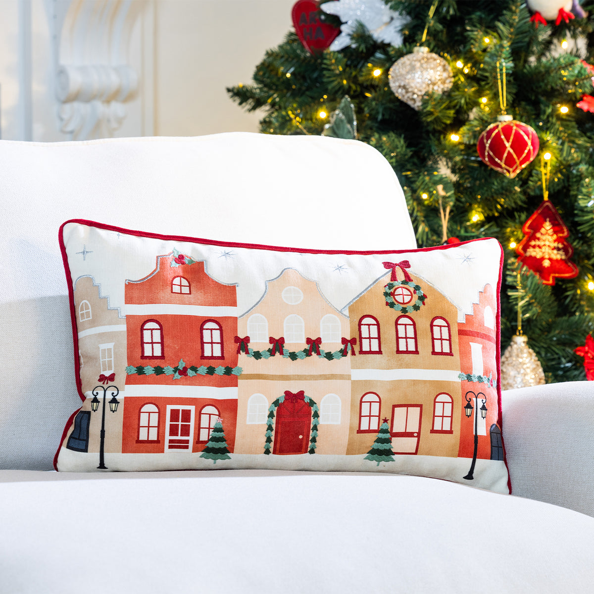Christmas Wishes Christmas Town Cushion featuring festive townhouses, displayed on a white sofa beside a decorated Christmas tree.