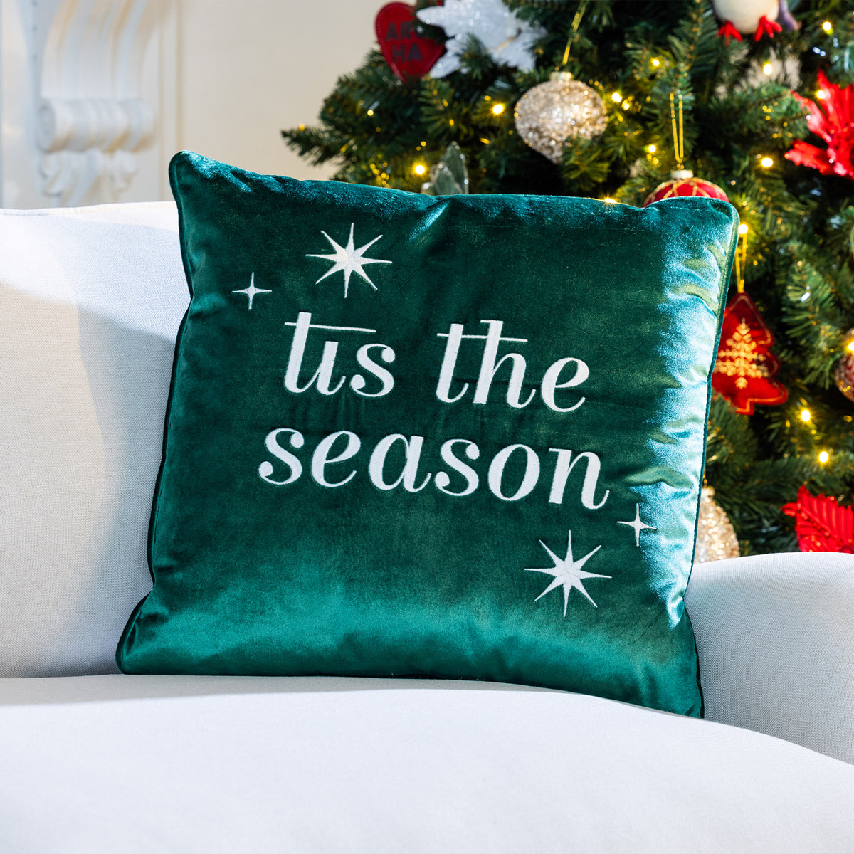 Green velvet 'Tis the Season Cushion featuring white embroidered lettering and stars, displayed on a white sofa beside a Christmas tree.