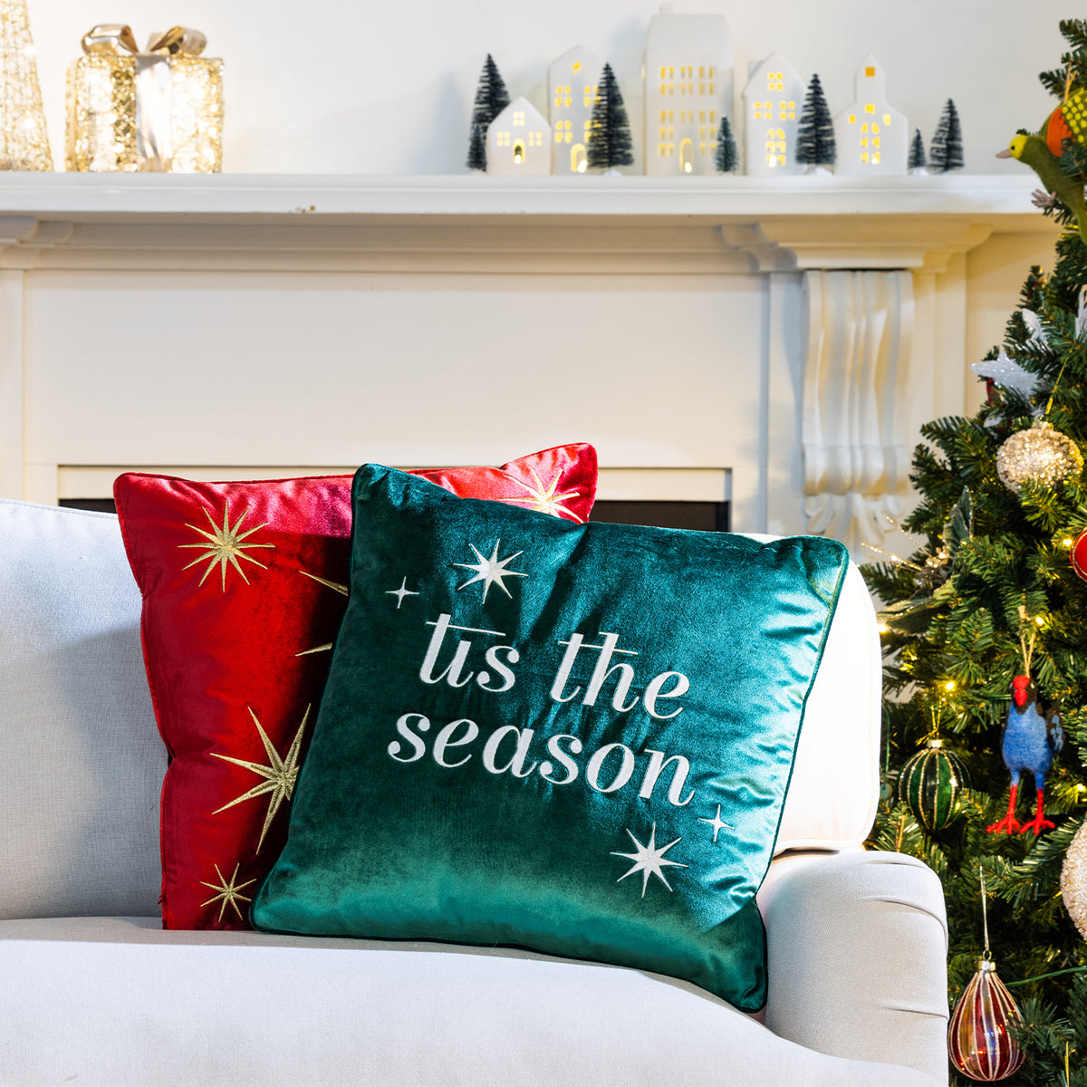 Red velvet Christmas Gold Star Cushion displayed behind a green 'Tis the Season Cushion on a white sofa beside a decorated Christmas tree.