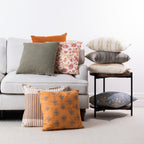 A styled collection of decorative cushions in various colors and patterns, arranged on a light gray sofa, on a small black side table, and on the floor against a white wall.