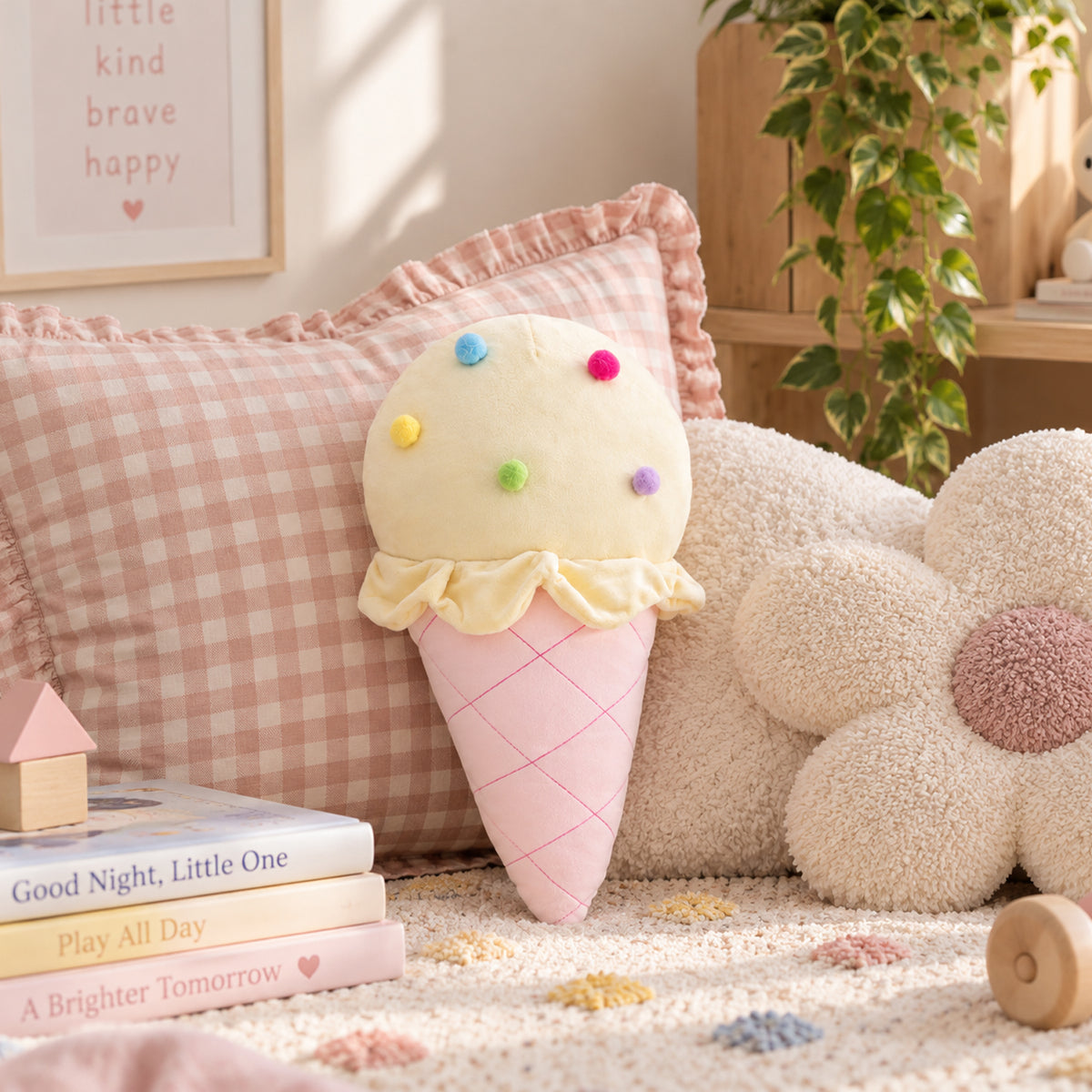 A plush ice cream cone cushion with a pale yellow scoop topped with colorful felt dots and a pink cone base, styled on a beige rug beside a pink gingham pillow, a flower-shaped cushion, and soft home decor props.