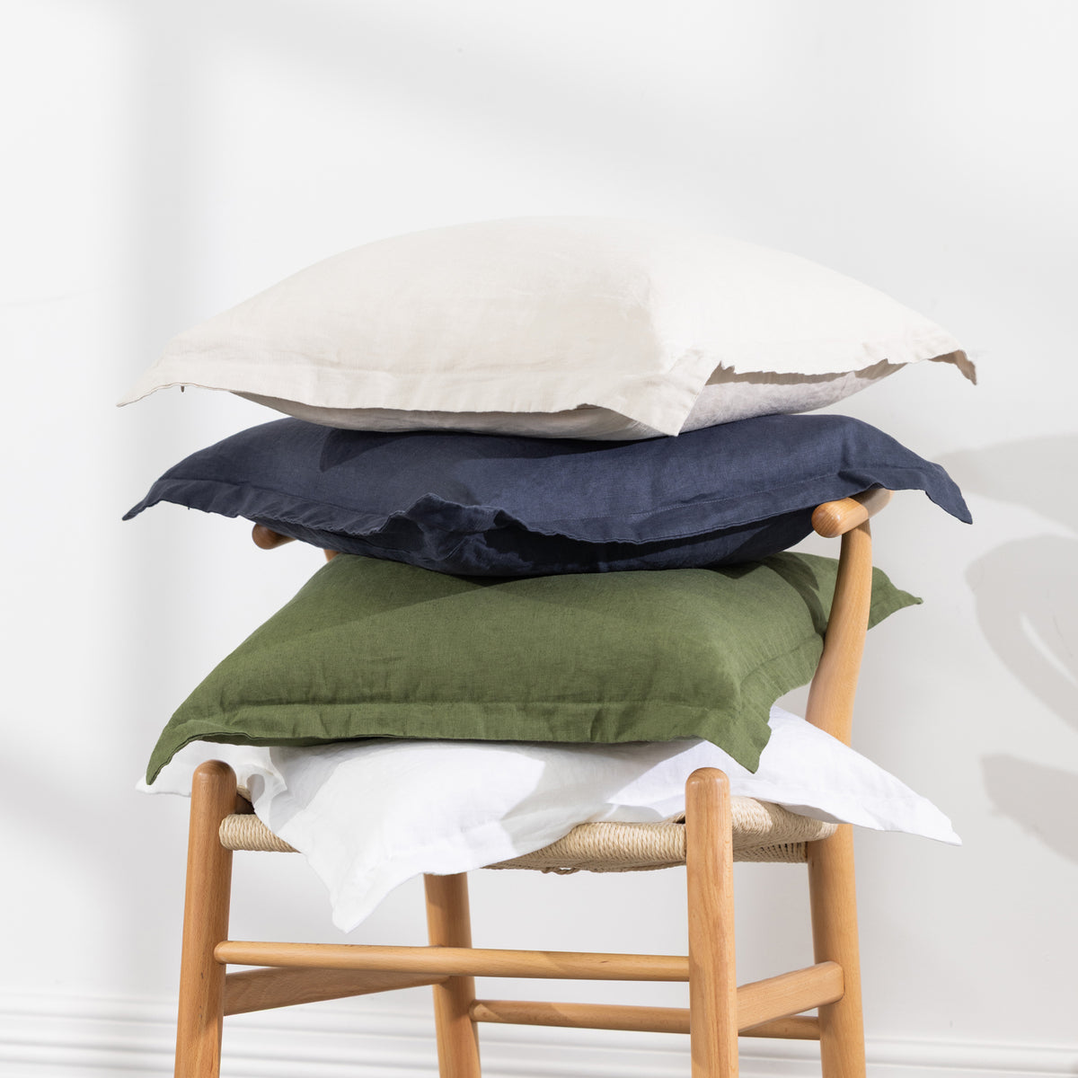 Four square throw pillows stacked on a wooden chair with a white sheet, set against a plain white wall.