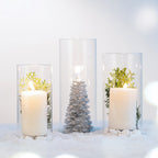 Decorative glass cylinder vases styled with candles, greenery, and festive accents on a snowy white surface.