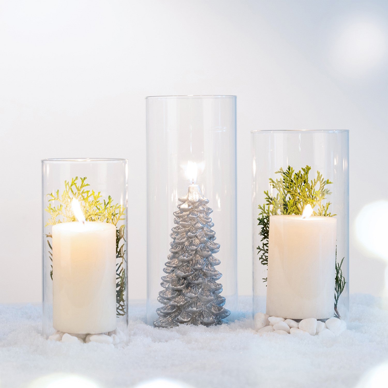 Decorative glass cylinder vases styled with candles, greenery, and festive accents on a snowy white surface.
