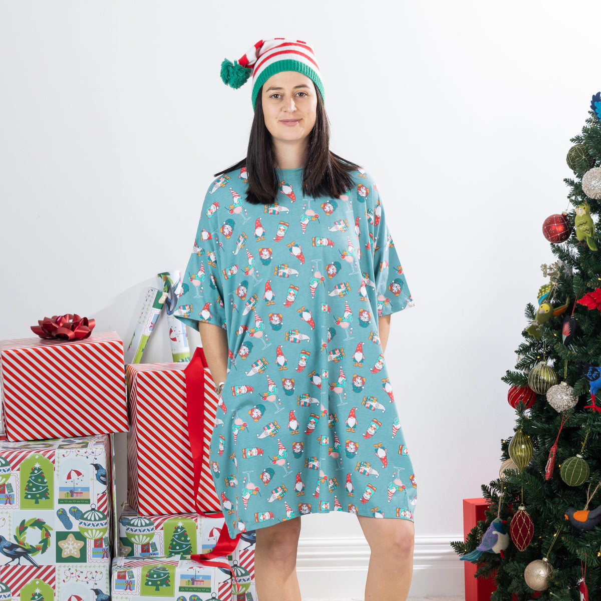 Woman wearing the Christmas Wishes Christmas Gnome Sleep Tee in jade with an all-over festive gnome print, styled with a striped Christmas hat beside wrapped presents and a decorated Christmas tree.