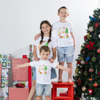 Three children wearing matching Christmas Wishes Cheer Kids Tees in white beside a decorated Christmas tree and wrapped presents.