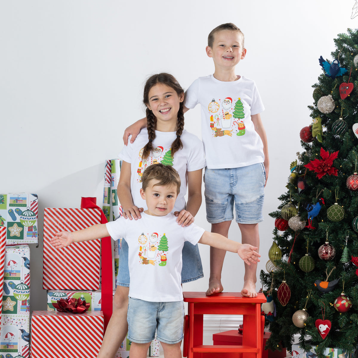 Three children wearing matching Christmas Wishes Cheer Kids Tees in white beside a decorated Christmas tree and wrapped presents.