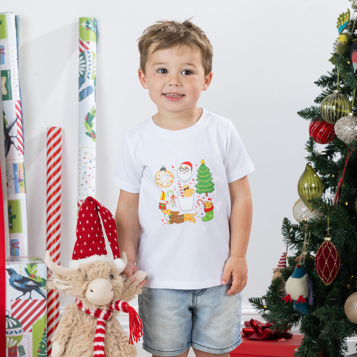 Young boy wearing the Christmas Wishes Cheer Kids Tee in white beside a Christmas tree and festive plush decoration.