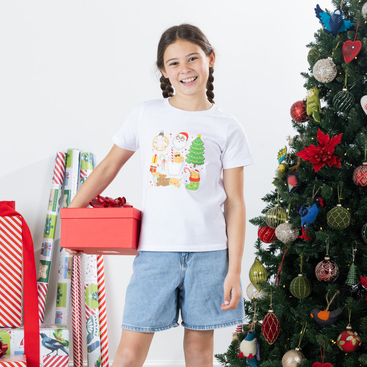 Girl wearing the Christmas Wishes Cheer Kids Tee in white while holding a red gift box beside a decorated Christmas tree.