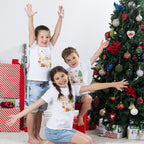 Three children wearing matching Christmas Wishes Cheer Kids Tees in white with arms raised in a playful festive Christmas scene.