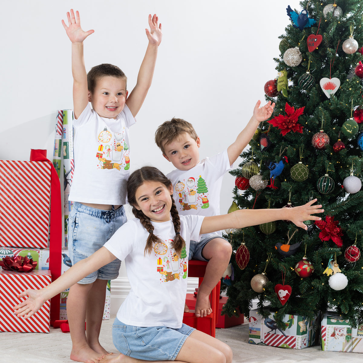 Three children wearing matching Christmas Wishes Cheer Kids Tees in white with arms raised in a playful festive Christmas scene.