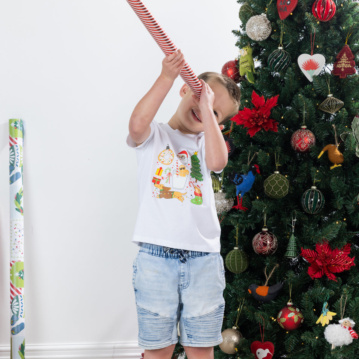 Boy wearing the Christmas Wishes Cheer Kids Tee in white while playfully looking through a roll of striped Christmas wrapping paper beside a decorated tree.