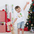 Young boy wearing the Christmas Wishes Cheer Kids Tee in white, posing beside wrapped presents and a decorated Christmas tree while holding a festive plush toy.