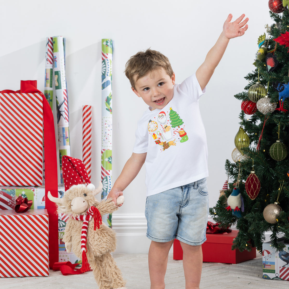 Young boy wearing the Christmas Wishes Cheer Kids Tee in white, posing beside wrapped presents and a decorated Christmas tree while holding a festive plush toy.