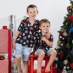 Two young boys wearing matching black Christmas Wishes pōhutukawa festive shirts beside a Christmas tree.