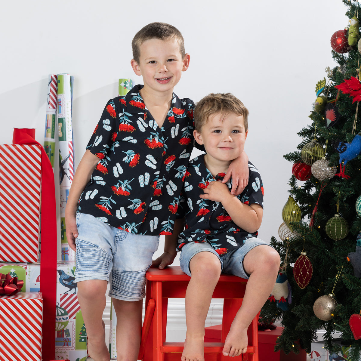 Two young boys wearing matching black Christmas Wishes pōhutukawa festive shirts beside a Christmas tree.
