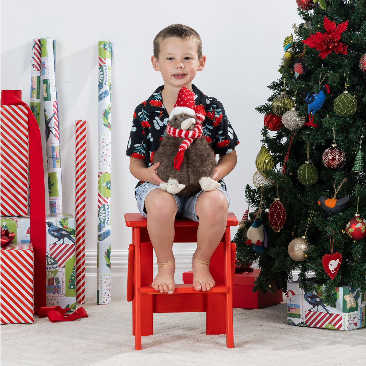Young boy wearing the black Christmas Wishes pōhutukawa festive shirt while seated with a Christmas plush toy.