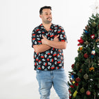 Man wearing the black Christmas Wishes pōhutukawa festive shirt with his arms crossed beside a Christmas tree.