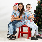 Three children wearing matching faded blue Christmas Wishes Cheeky Santa festive shirts beside a Christmas tree.