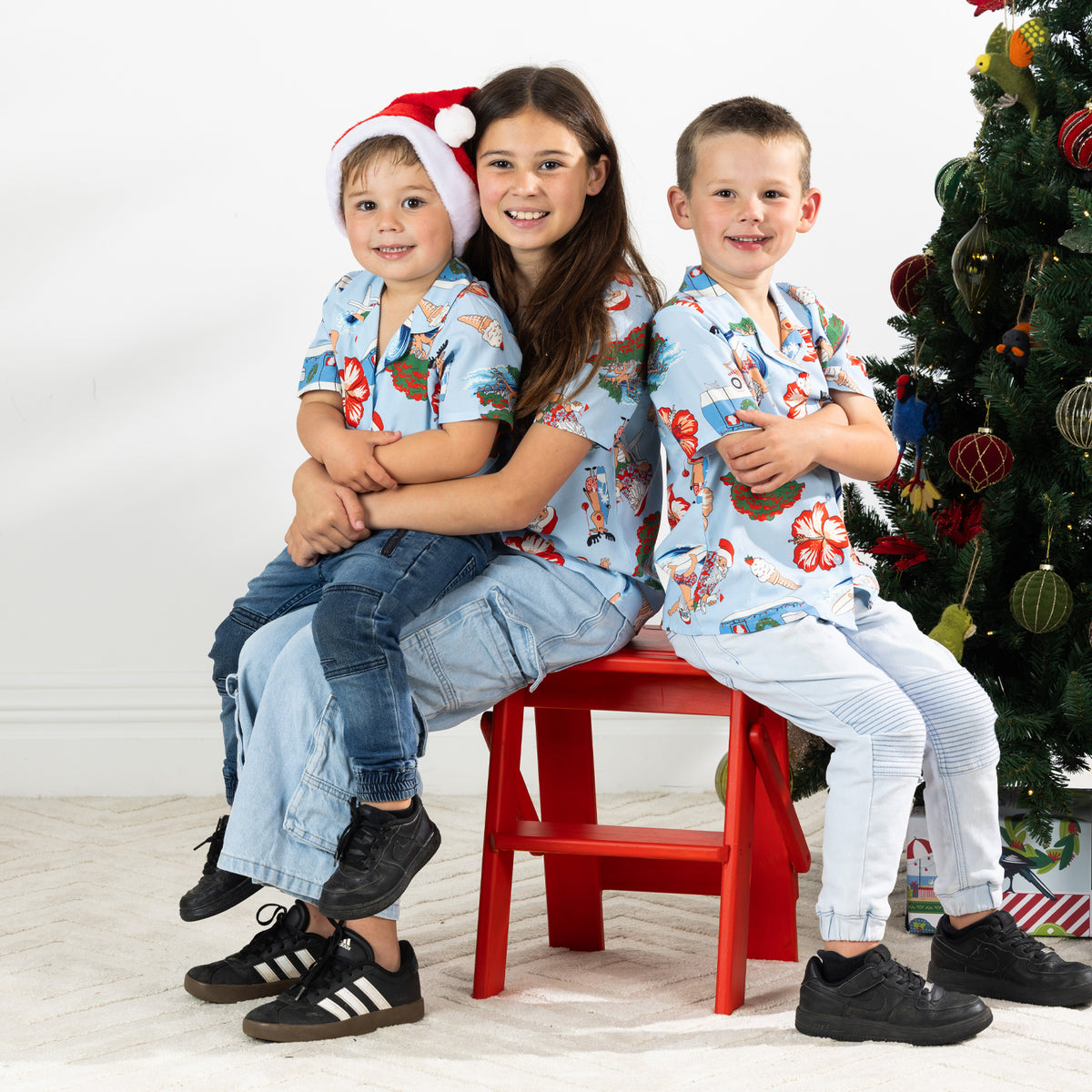 Three children wearing matching faded blue Christmas Wishes Cheeky Santa festive shirts beside a Christmas tree.