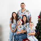 Family wearing matching faded blue Christmas Wishes Cheeky Santa festive shirts beside a decorated Christmas tree.