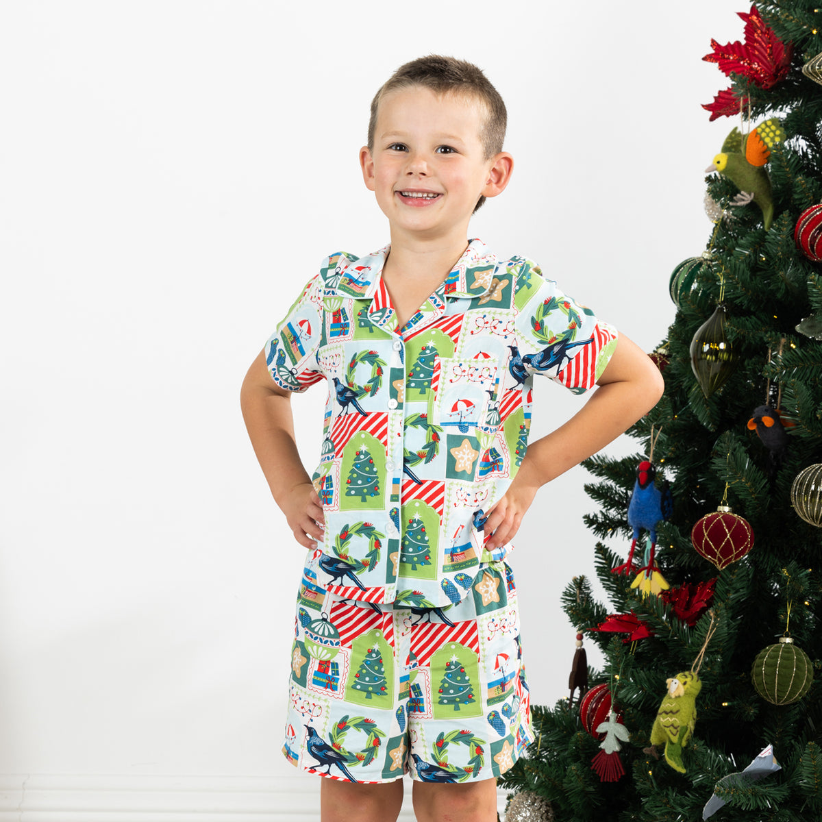 Boy wearing the Christmas Wishes Tui Kids PJ Set in white beside a decorated Christmas tree.