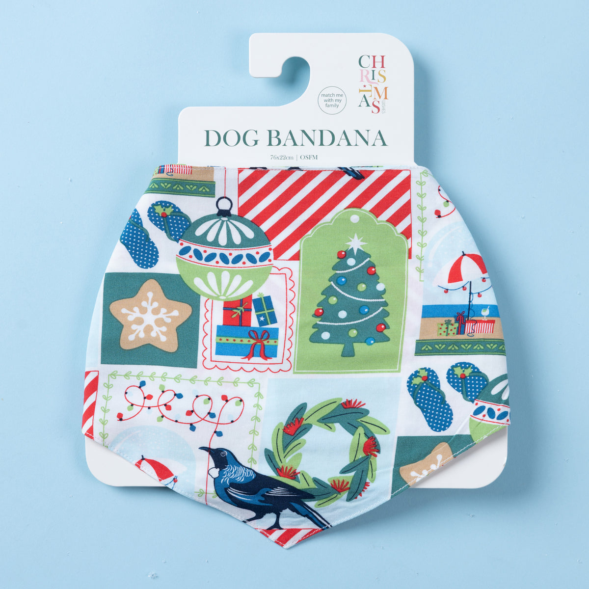 White Christmas Wishes dog bandana displayed on branded packaging, featuring Christmas trees, ornaments, gifts, wreaths, jandals and tui birds.