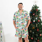 Man wearing the Christmas Wishes Tui Men’s PJ Set in white with a button-up short sleeve top and matching shorts beside a decorated Christmas tree.