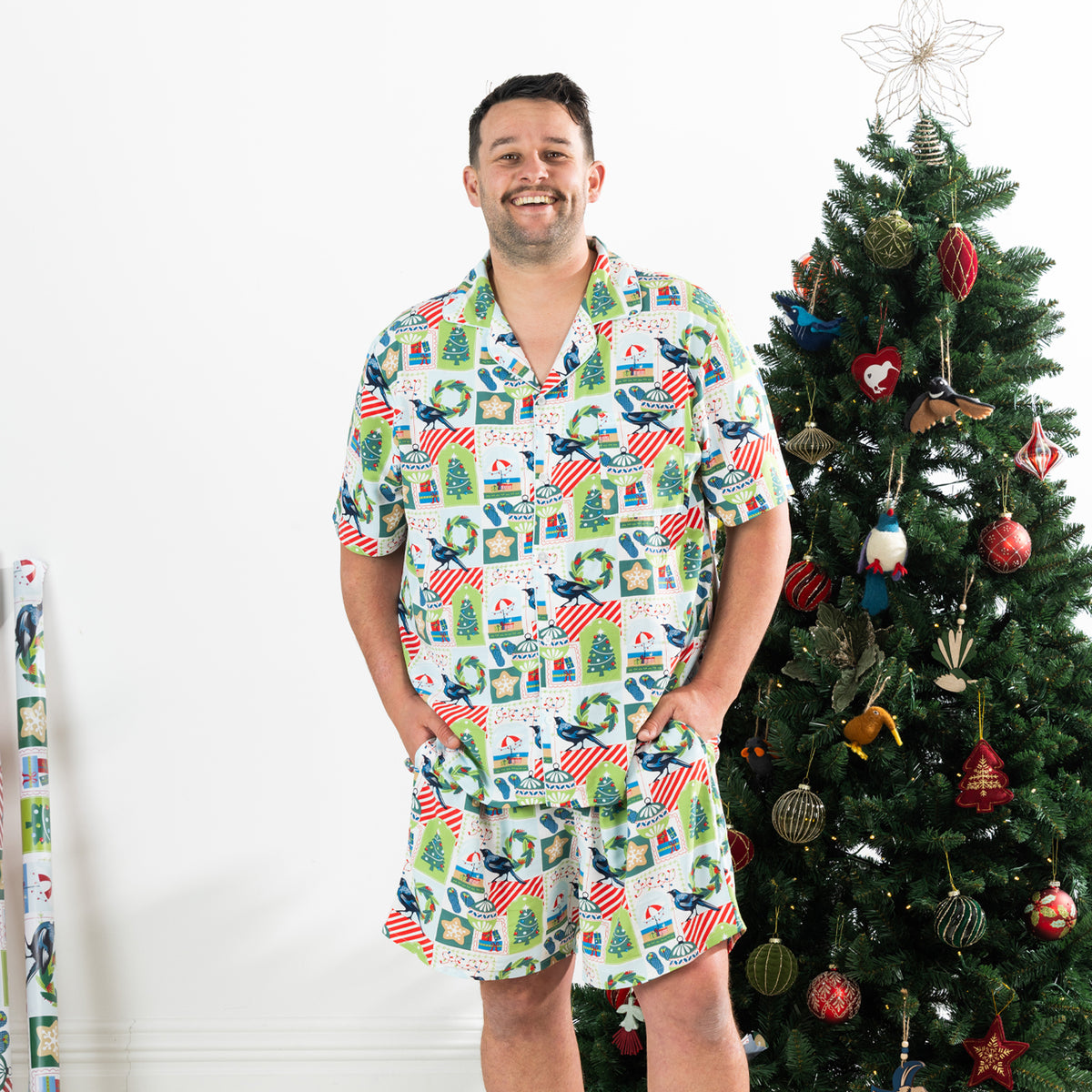 Man wearing the Christmas Wishes Tui Men’s PJ Set in white with a button-up short sleeve top and matching shorts beside a decorated Christmas tree.