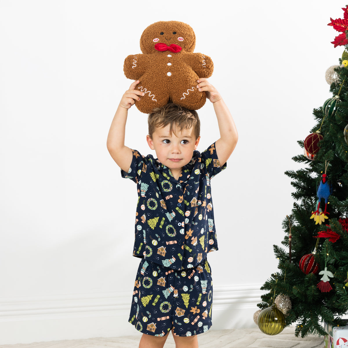 Boy wearing the Christmas Wishes Wreath Kids PJ Set in navy while holding a gingerbread cushion above his head.