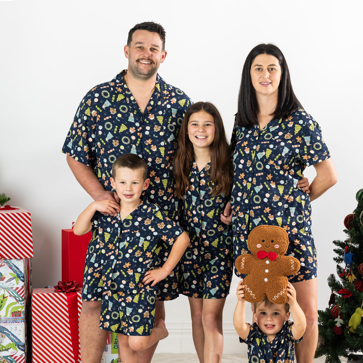 Family wearing matching Christmas Wishes Wreath PJ Sets in navy beside a decorated Christmas tree.