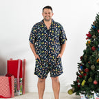 Man wearing the Christmas Wishes Wreath Men’s PJ Set in navy with hands in his pockets beside a decorated Christmas tree.
