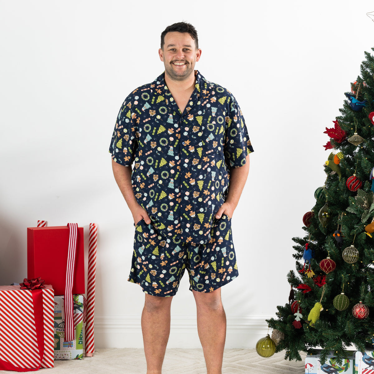 Man wearing the Christmas Wishes Wreath Men’s PJ Set in navy with hands in his pockets beside a decorated Christmas tree.