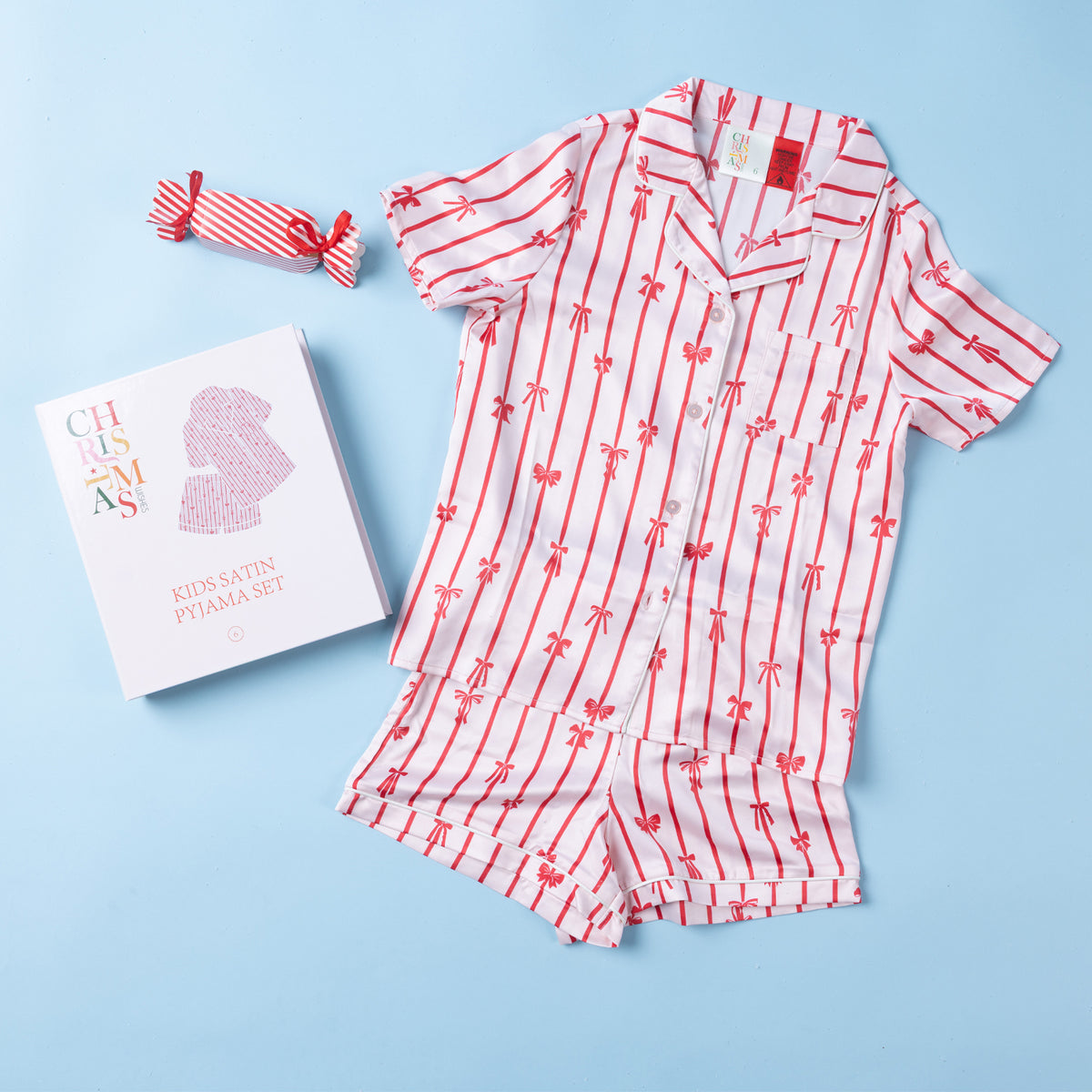 Christmas Wishes Kids’ Satin Bow PJ Set displayed with matching gift box and striped cracker-style accessory on a light blue background.