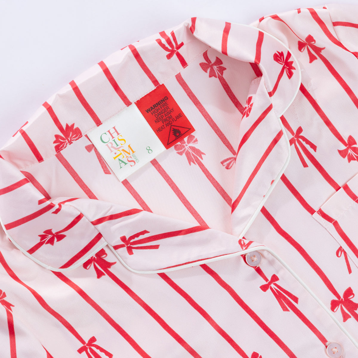 Close-up of the Christmas Wishes Kids’ Satin Bow PJ Set showing the red striped bow print, piped collar, button front and warning label.