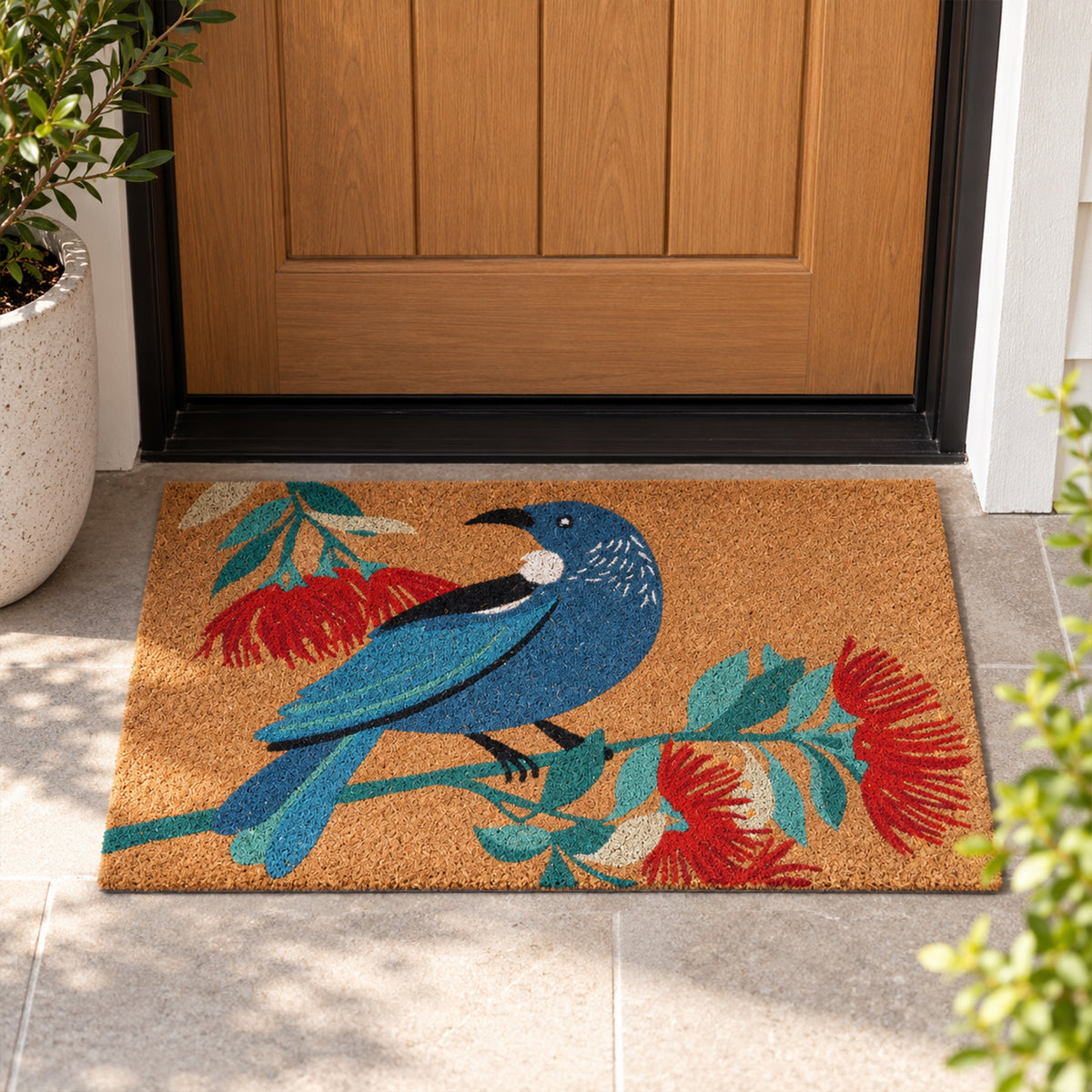 Christmas Wishes Xmas Tui Coir Mat featuring a blue tūī perched on a branch surrounded by red pōhutukawa flowers and green foliage against a natural coir background.