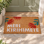 Merry Kirihimete Coir Mat displayed on a tiled doorstep in front of a sage green door, surrounded by greenery in natural sunlight.