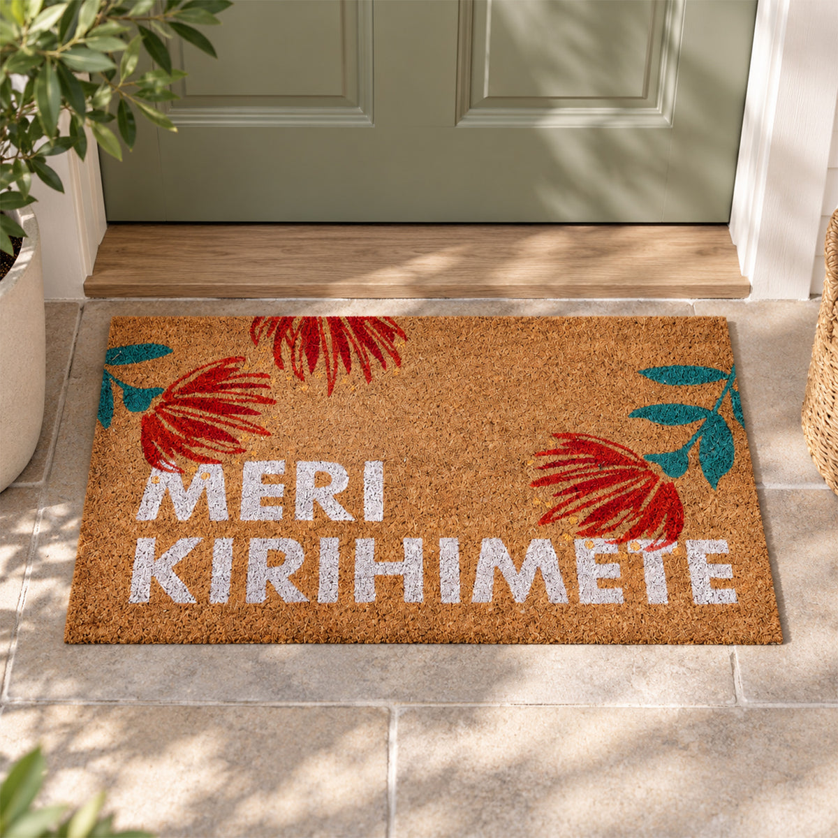 Merry Kirihimete Coir Mat displayed on a tiled doorstep in front of a sage green door, surrounded by greenery in natural sunlight.