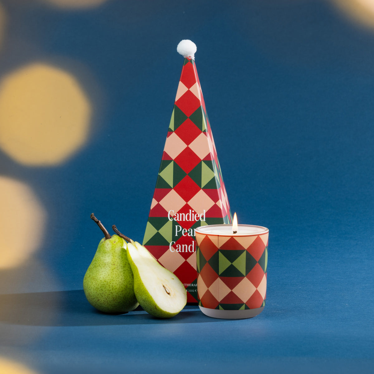 A festive Candied Pear candle with bold geometric Christmas packaging, styled alongside fresh pears against a rich blue backdrop.