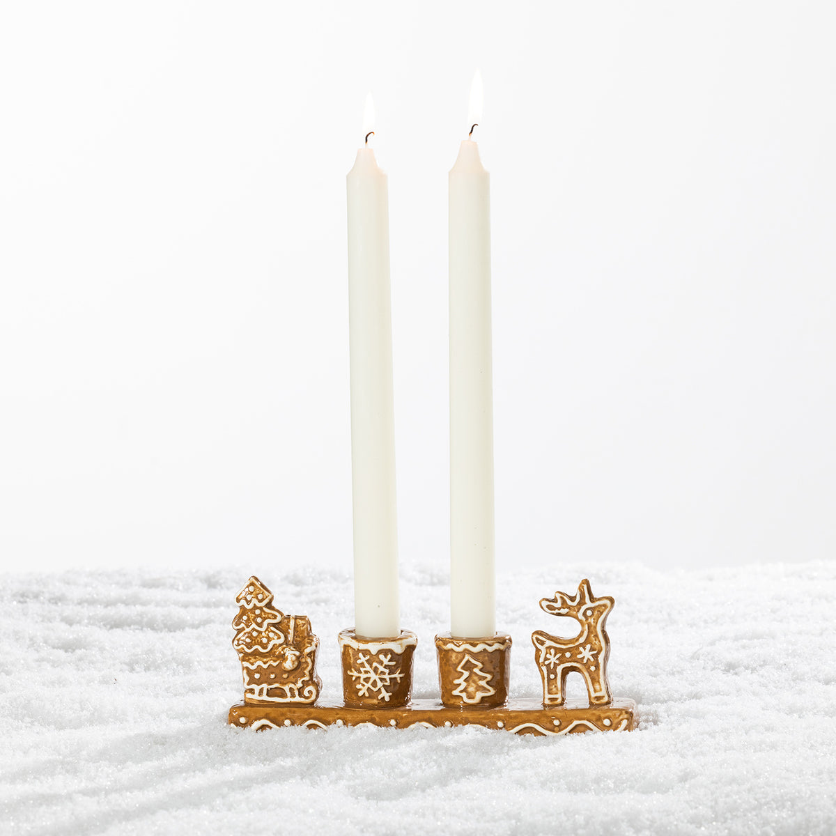 Gingerbread-style ceramic two-candle holder displayed with two tall white taper candles on a snowy festive background.