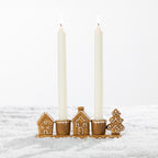 Gingerbread-style ceramic two-candle holder displayed with two tall white taper candles, featuring festive houses and a Christmas tree on a snowy background.