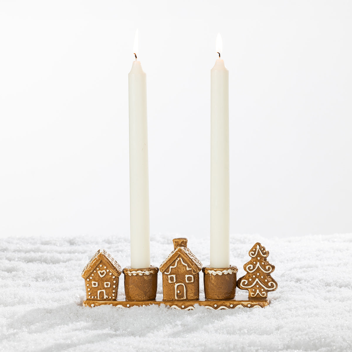 Gingerbread-style ceramic two-candle holder displayed with two tall white taper candles, featuring festive houses and a Christmas tree on a snowy background.