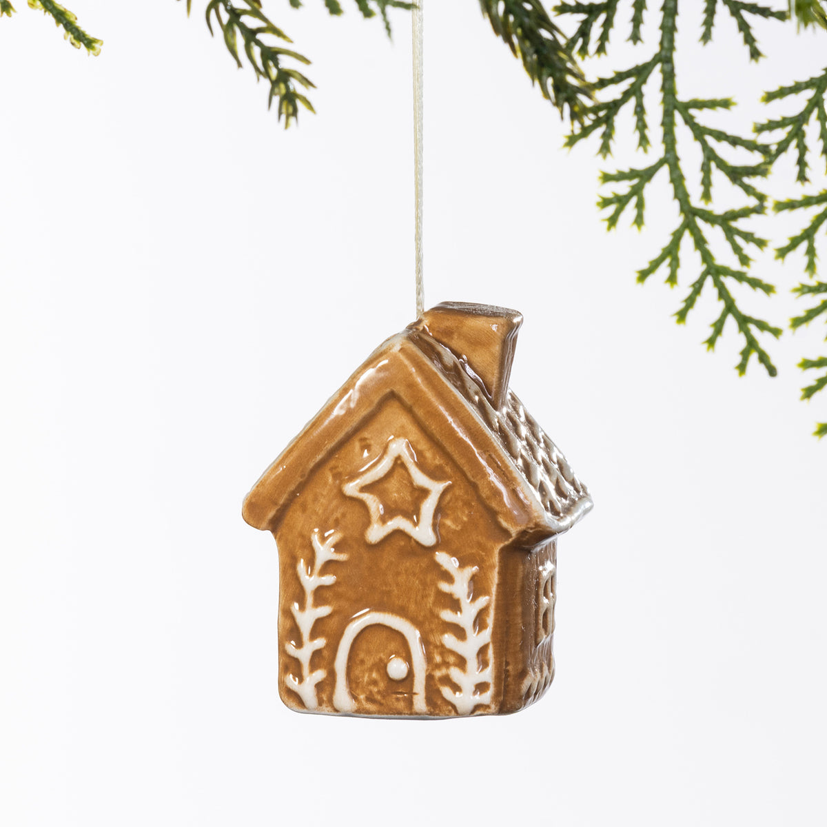 Gingerbread-style ceramic house hanging decoration with white star and branch detailing, displayed hanging from a Christmas tree branch.