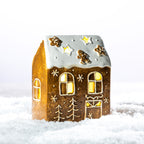 Gingerbread-style ceramic LED house viewed from an angle, featuring a snowy white roof, festive detailing and warm glowing windows.