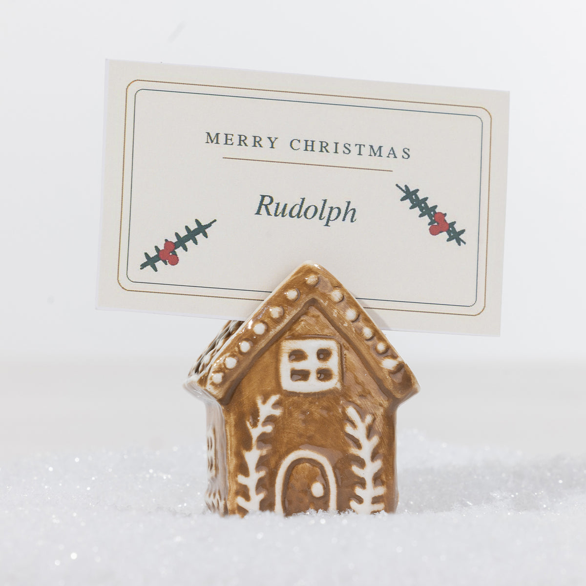 Gingerbread-style ceramic house name card holder with white icing details, holding a personalised Merry Christmas place card in a snowy setting.