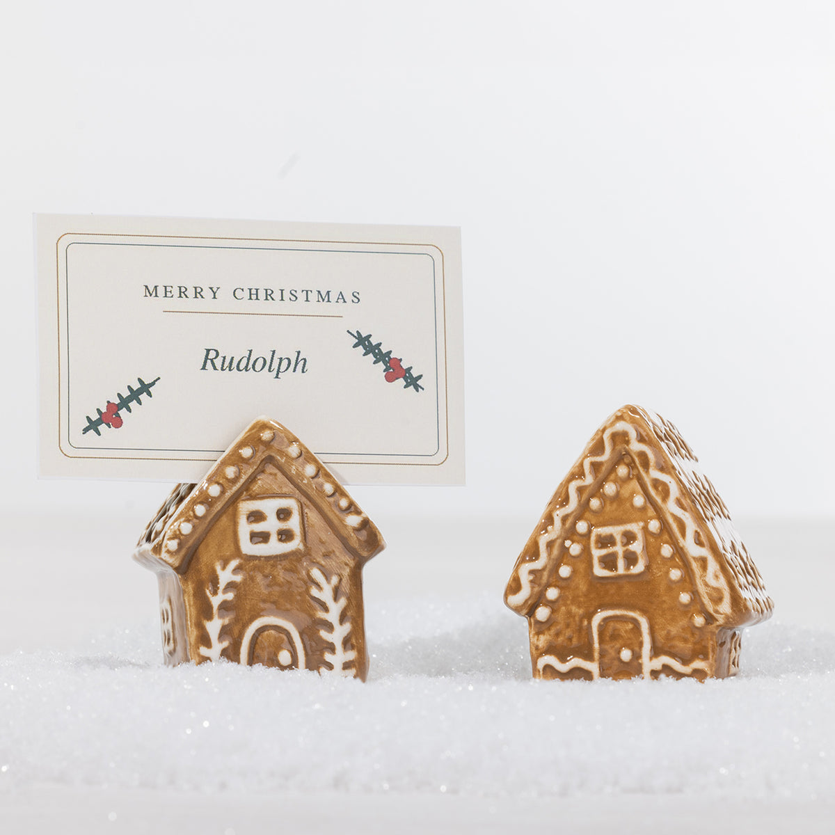 Two gingerbread-style ceramic house name card holders displayed in snow, with one holding a personalised Merry Christmas place card.