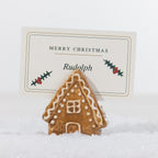 Gingerbread-style ceramic house name card holder with white icing details, holding a personalised Merry Christmas place card in a snowy setting.